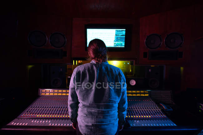 Vista posteriore di un giovane ingegnere del suono caucasico in piedi su un banco di miscelazione in uno studio di registrazione a guardare un monitor del computer — Foto stock