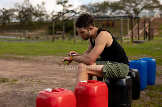 Вид збоку молодого Кавказького чоловіка сидить і перевіряючи свій годинник на відкритому спортзалі перед Bootcamp тренувальна сесія — стокове фото