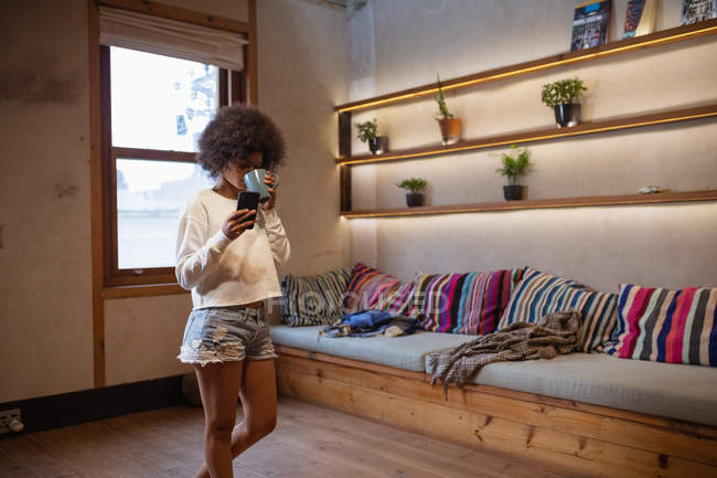 Vista lateral de uma jovem mulher de raça mista em pé em sua sala de estar, usando um smartphone e bebendo uma xícara de café — Fotografia de Stock