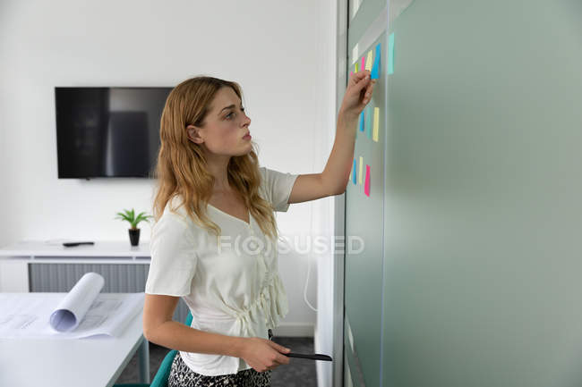 Vista lateral de cerca de una joven mujer caucásica de pie y pegando notas adhesivas de colores en una pared de vidrio en la oficina moderna de un negocio creativo - foto de stock
