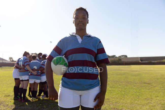 Portrait d'une jeune joueuse de rugby mixte adulte debout sur un terrain de rugby avec une balle de rugby sous son bras regardant vers la caméra, avec ses coéquipières dans un câlin ensemble en arrière-plan — Photo de stock