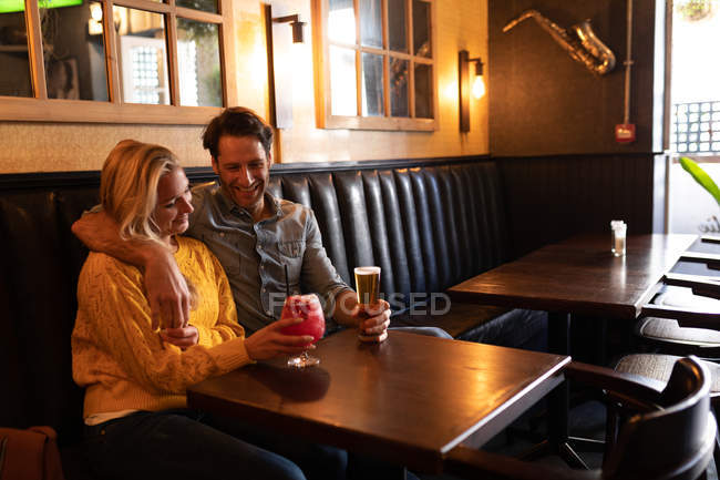 Vista lateral de una feliz pareja caucásica joven relajándose juntos en vacaciones en un bar, abrazando, bebiendo cerveza y cóctel - foto de stock