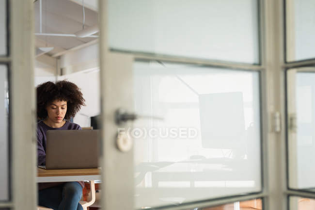 Frontansicht einer jungen Frau mit gemischter Rasse, die in einem kreativen Büro mit Laptop am Schreibtisch sitzt — Stockfoto