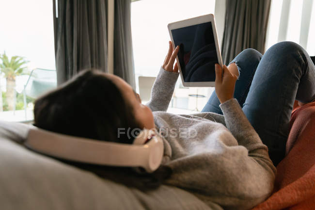 Над плечем зору молодий Кавказький жінка брюнетка лежачи на дивані з ногами вгору, носіння навушників і дивитися планшетний комп'ютер — стокове фото