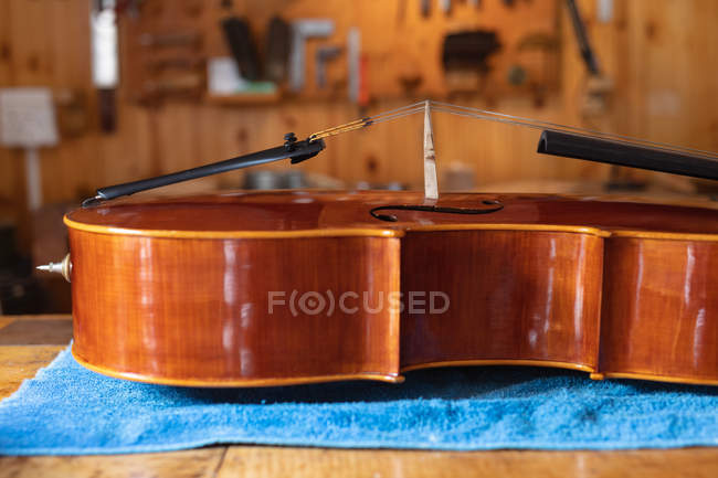 Primer plano de un violonchelo sobre un paño azul en un taller con herramientas colgando de la pared en el fondo - foto de stock