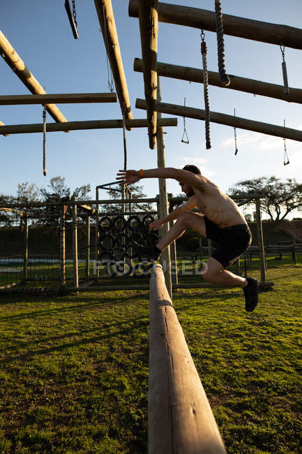 Vue latérale d'un jeune homme caucasien torse nu sautant par-dessus un obstacle dans une salle de gym extérieure lors d'une séance d'entraînement bootcamp — Photo de stock