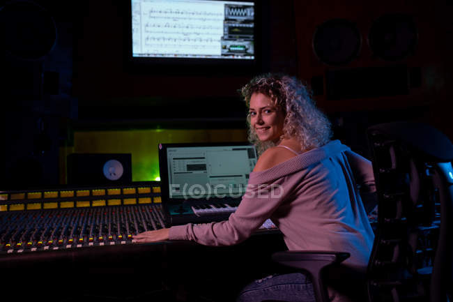 Vista trasera de una joven ingeniera de sonido caucásica sentada y trabajando en una mesa de mezclas en un estudio de grabación girando y sonriendo a la cámara - foto de stock