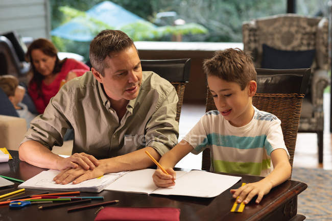 Front view of a middle aged Caucasian man helping his pre teen son with homework, the mother talking with their other son in the background — Stock Photo