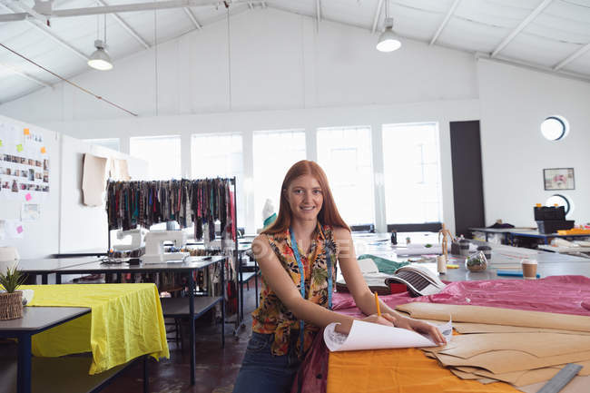 Porträt einer jungen kaukasischen Modestudentin, die in die Kamera lächelt, während sie in einem Atelier der Modehochschule an einem Design arbeitet — Stockfoto