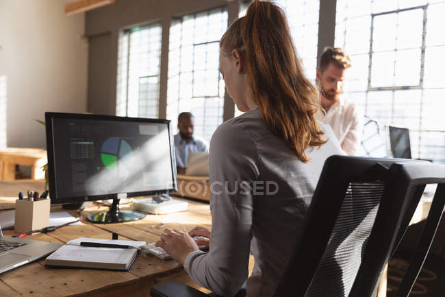 Vista trasera de cerca de una joven mujer caucásica sentada en un escritorio usando una computadora en una oficina creativa, con dos colegas masculinos sentados y trabajando en el fondo - foto de stock
