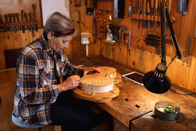 Vista lateral de una mujer luthier caucásica mayor trabajando en el cuerpo de un violín con una tableta en un banco de trabajo y herramientas colgando en la pared en el fondo - foto de stock