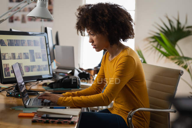 Vista laterale da vicino di una giovane donna di razza mista seduta a una scrivania utilizzando un computer portatile in un ufficio creativo — Foto stock