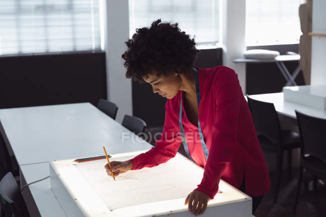 Side view of a young mixed race female fashion student working on a design drawing on a lightbox in a studio at fashion college — Stock Photo