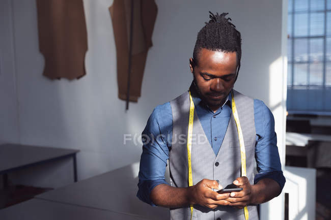 Vista frontal de un joven afroamericano estudiante de moda con una cinta métrica alrededor del cuello usando un teléfono inteligente en un estudio de la universidad de moda - foto de stock