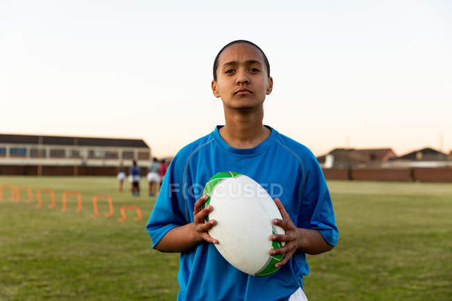 Ritratto di una giovane giocatrice di rugby di razza mista in piedi su un campo sportivo che tiene una palla da rugby durante una sessione di allenamento — Foto stock