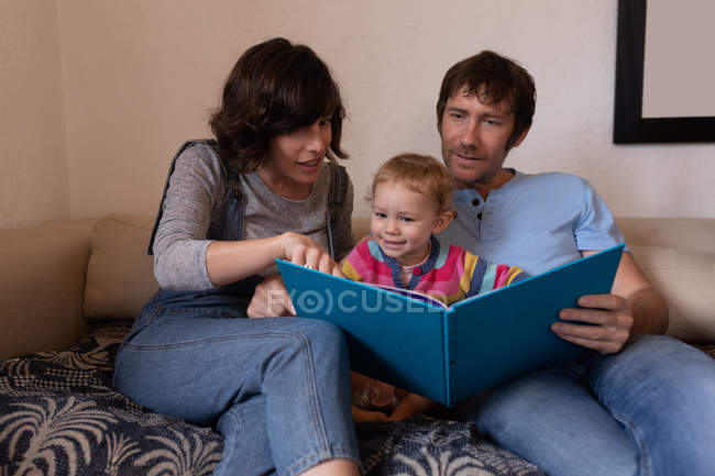 Обзор молодых кавказских родителей, сидящих на диване и читающих книгу со своим ребенком — стоковое фото