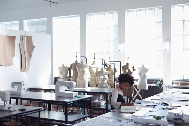 Vista lateral de una joven estudiante de moda de raza mixta sentada en una mesa trabajando en un diseño en un estudio de la universidad de moda con maniquíes en el fondo - foto de stock