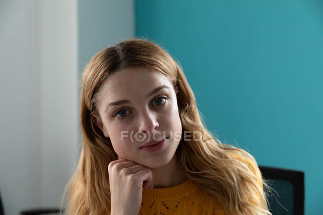 Portrait close up of a young Caucasian woman with long blonde hair and blue eyes resting her chin on her hand, looking straight to camera in the office of a creative business — Stock Photo