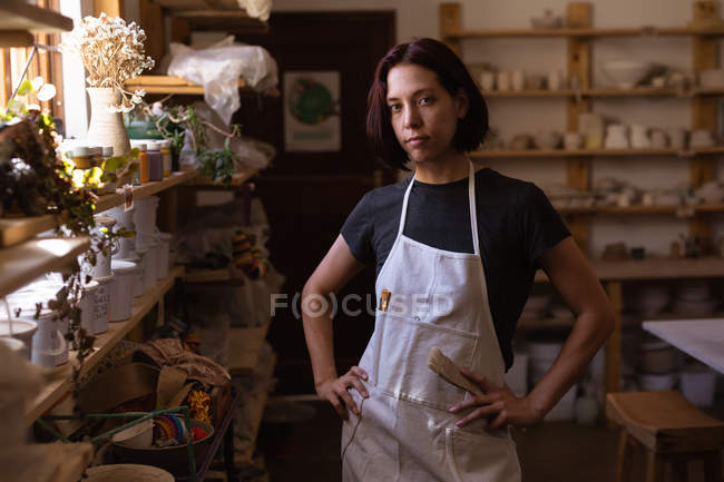 Портрет молодой кавказской женщины-гончара, стоящей с руками на бедрах в керамической мастерской, смотрящей в камеру — стоковое фото