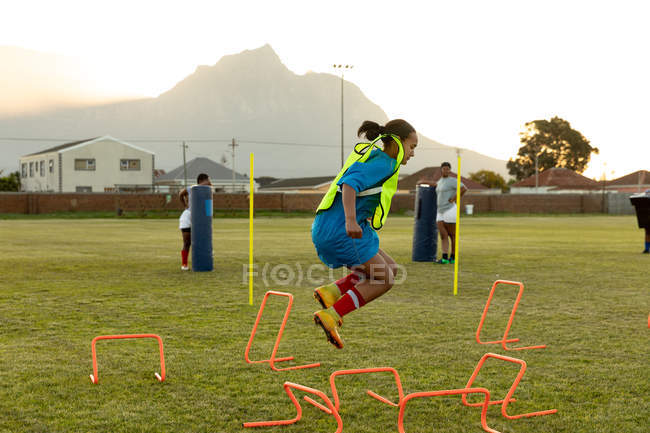 Side view of a young adult mixed race female rugby player jumping over agility hurdles on a rugby field during a training session, with teammates in the background — Stock Photo