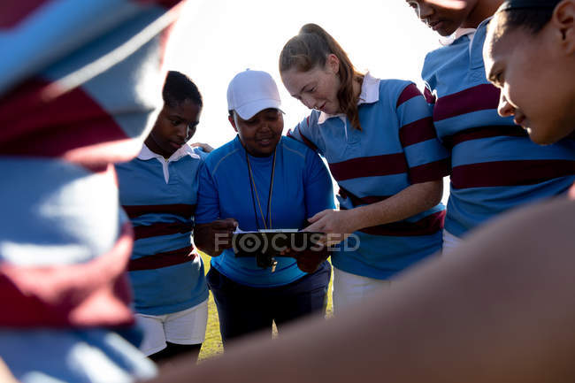 Frontansicht einer Mannschaft junger erwachsener multiethnischer Rugbyspielerinnen, die auf einem Rugbyfeld stehen, die Arme in einem Gedränge verbunden, während sie ihrer Trainerin mittleren Alters bei der Vorbereitung auf ein Rugbyspiel zuhören — Stockfoto