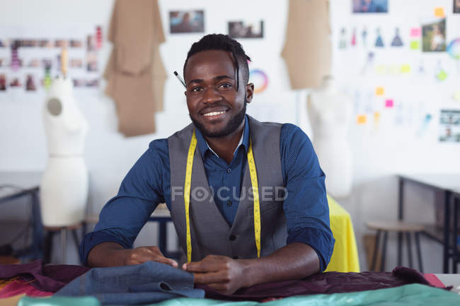 Портрет молодого афроамериканского студента-модельера с рулеткой на шее, улыбающегося в камеру и работающего над дизайном в студии колледжа моды — стоковое фото