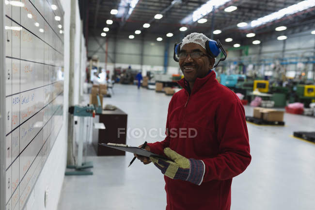Вигляд збоку закритий середнього віку Афроамериканський робітник на фабриці, який тримає на дошці перевірку на дошці, звертаючись до камери і посміхаючись на складі на заводі обробки. — стокове фото