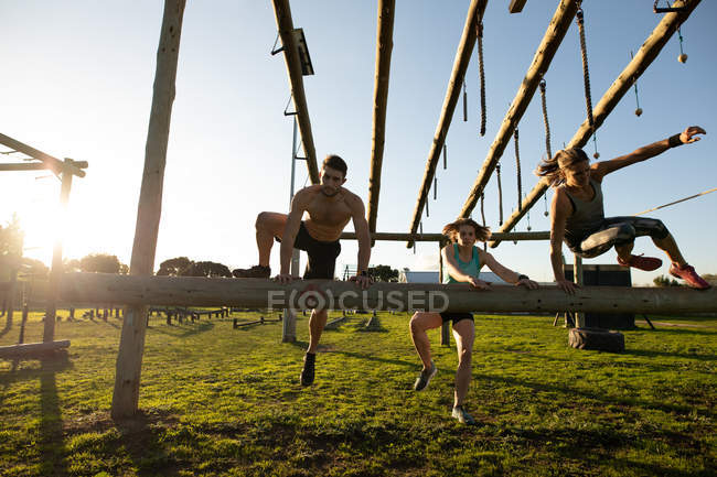 Vista frontal de dos jóvenes mujeres caucásicas y un joven hombre caucásico saltando sobre un obstáculo en un gimnasio al aire libre durante una sesión de entrenamiento de bootcamp - foto de stock