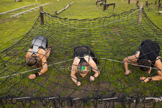 Front view close up of two young Caucasian women and a young Caucasian man crawling under a net at an outdoor gym during a bootcamp training session — Stock Photo
