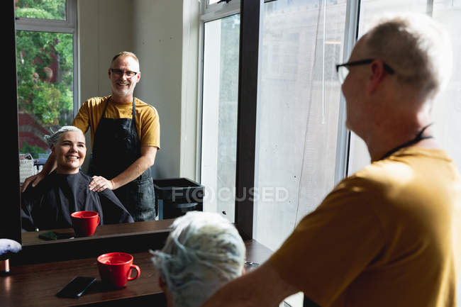 Rear view of a middle aged Caucasian male hairdresser and a young Caucasian woman having her hair styled in a hair salon, reflected in a mirror — Stock Photo