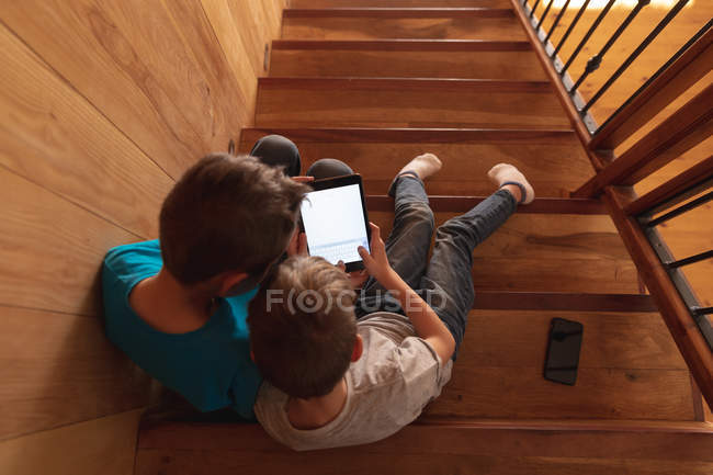 Вид сверху на двух кавказских мальчиков, сидящих дома на лестнице, с помощью планшетного компьютера и смартфона — стоковое фото