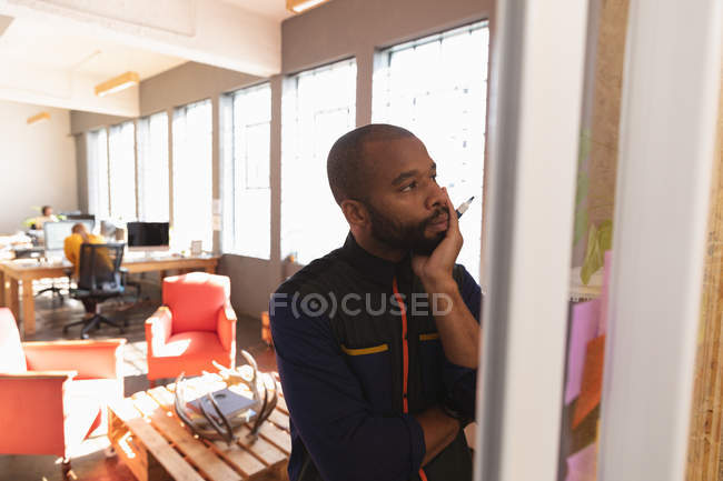Vista frontal close-up de um jovem afro-americano lendo notas em uma parede e pensando durante uma sessão de brainstorm em equipe em um escritório criativo — Fotografia de Stock