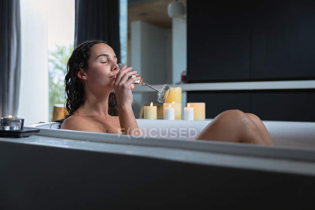 Vista lateral de una joven morena caucásica sentada en un baño con velas encendidas al lado, bebiendo champán con los ojos cerrados - foto de stock