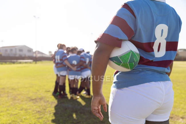 Rückansicht Mittelteil einer jungen erwachsenen Mixed-Race-Rugbyspielerin, die mit einem Rugbyball unter dem Arm auf einem Rugbyfeld steht, während sich ihre Teamkolleginnen im Hintergrund eng aneinander drängen — Stockfoto
