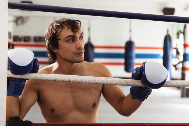 Vue de face gros plan d'un jeune boxeur masculin mixte appuyé sur une corde d'un ring de boxe — Photo de stock