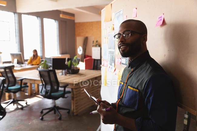 Vue de côté gros plan d'un jeune homme afro-américain souriant utilisant une tablette informatique debout près d'un tableau d'humeur lors d'une séance de remue-méninges d'équipe dans un bureau créatif, avec des collègues travaillant en arrière-plan — Photo de stock