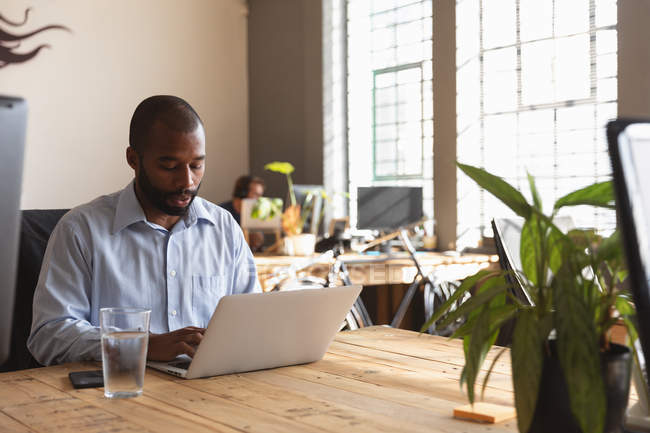 Vista frontal de perto de um jovem afro-americano sentado em uma mesa com um copo de água usando um computador portátil em um escritório criativo, com um colega trabalhando em segundo plano — Fotografia de Stock