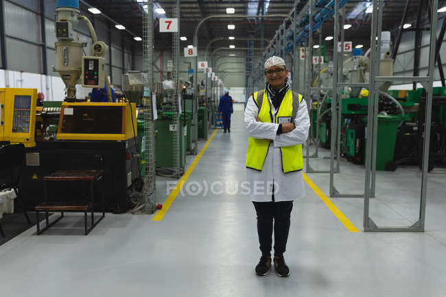 Ritratto di una donna caucasica di mezza età che indossa occhiali e abbigliamento da lavoro in piedi tra file di attrezzature sorridenti in un magazzino presso uno stabilimento di lavorazione, un altro lavoratore visibile sullo sfondo — Foto stock
