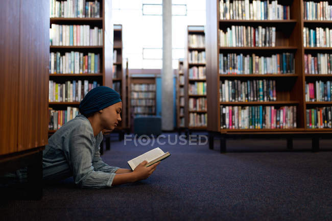 Vista lateral de uma jovem estudante asiática vestindo um turbante lendo um livro, deitada em um chão e estudando em uma biblioteca — Fotografia de Stock