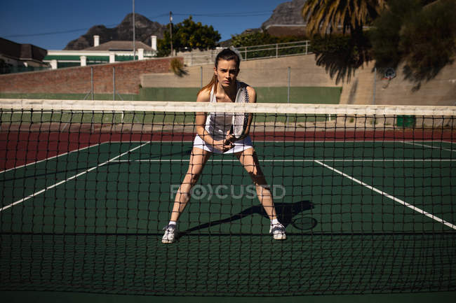 Vorderansicht einer jungen kaukasischen Frau, die an einem sonnigen Tag Tennis spielt, einen Schläger in der Hand hält und auf den Ball wartet, durch das Netz gesehen — Stockfoto