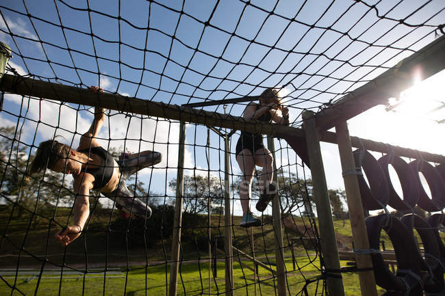 Vista frontal de duas jovens mulheres caucasianas escalando redes em uma estrutura de escalada em um ginásio ao ar livre durante uma sessão de treinamento de bootcamp — Fotografia de Stock