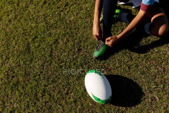Sección baja de alto ángulo de la jugadora de rugby sentada y atando su bota en un campo de rugby, con la pelota a su lado - foto de stock