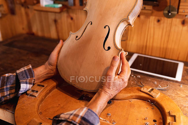 Primer plano de la luthier femenina sosteniendo el cuerpo de un violín con una tableta en un banco de trabajo - foto de stock