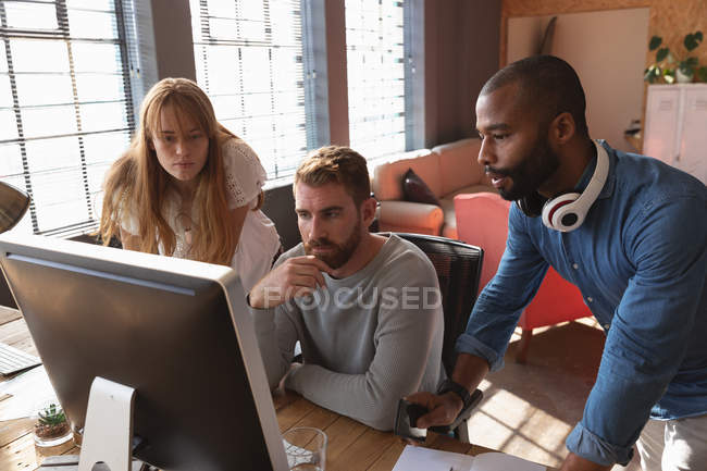 Vue de face gros plan d'un jeune homme afro-américain et d'une jeune femme caucasienne debout de chaque côté d'un jeune collègue caucasien assis à un bureau à l'aide d'un ordinateur, regardant son moniteur ensemble et parlant dans un bureau créatif — Photo de stock