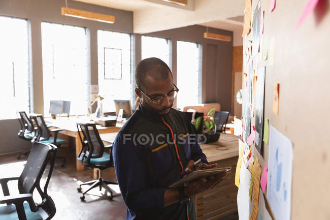 Vista frontal close-up de um jovem afro-americano usando um computador tablet em pé por um quadro de humor durante uma sessão de brainstorm em equipe em um escritório criativo — Fotografia de Stock