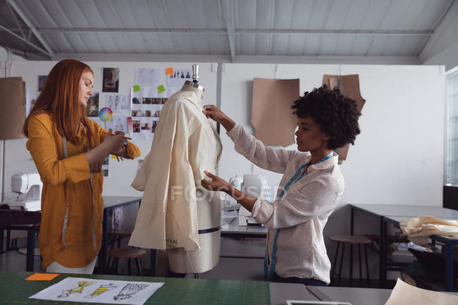 Vue de côté d'une jeune afro-américaine et d'une jeune étudiante caucasienne travaillant ensemble sur la conception d'un vêtement sur un mannequin dans un studio au collège de mode — Photo de stock