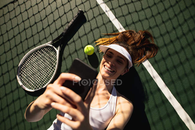 Ansicht einer jungen kaukasischen Frau, die an einem sonnigen Tag auf einem Tennisplatz liegt und ein Selfie macht — Stockfoto