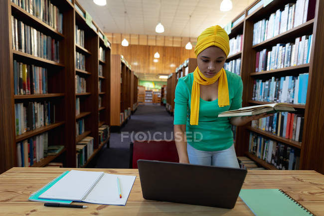 Передній погляд на молоду азіатську студентку, одягнену в хіджаб, що тримає книжку, за допомогою ноутбука і навчаючись у бібліотеці. — стокове фото
