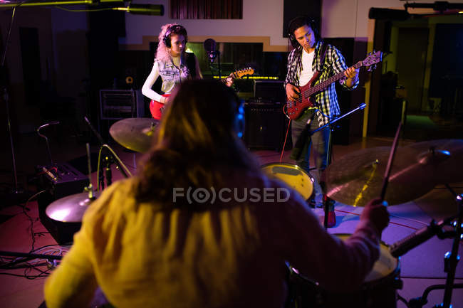 Rear view close up of a young Caucasian male drummer playing a drum kit with a young Caucasian woman wearing headphones playing electric guitar and a young mixed race man wearing headphones playing bass guitar facing him — Stock Photo