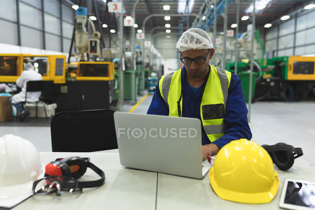 Front view close up of a middle aged mixed race male factory worker wearing glasses and workwear using a laptop computer in a warehouse at a factory processing plant — Stock Photo